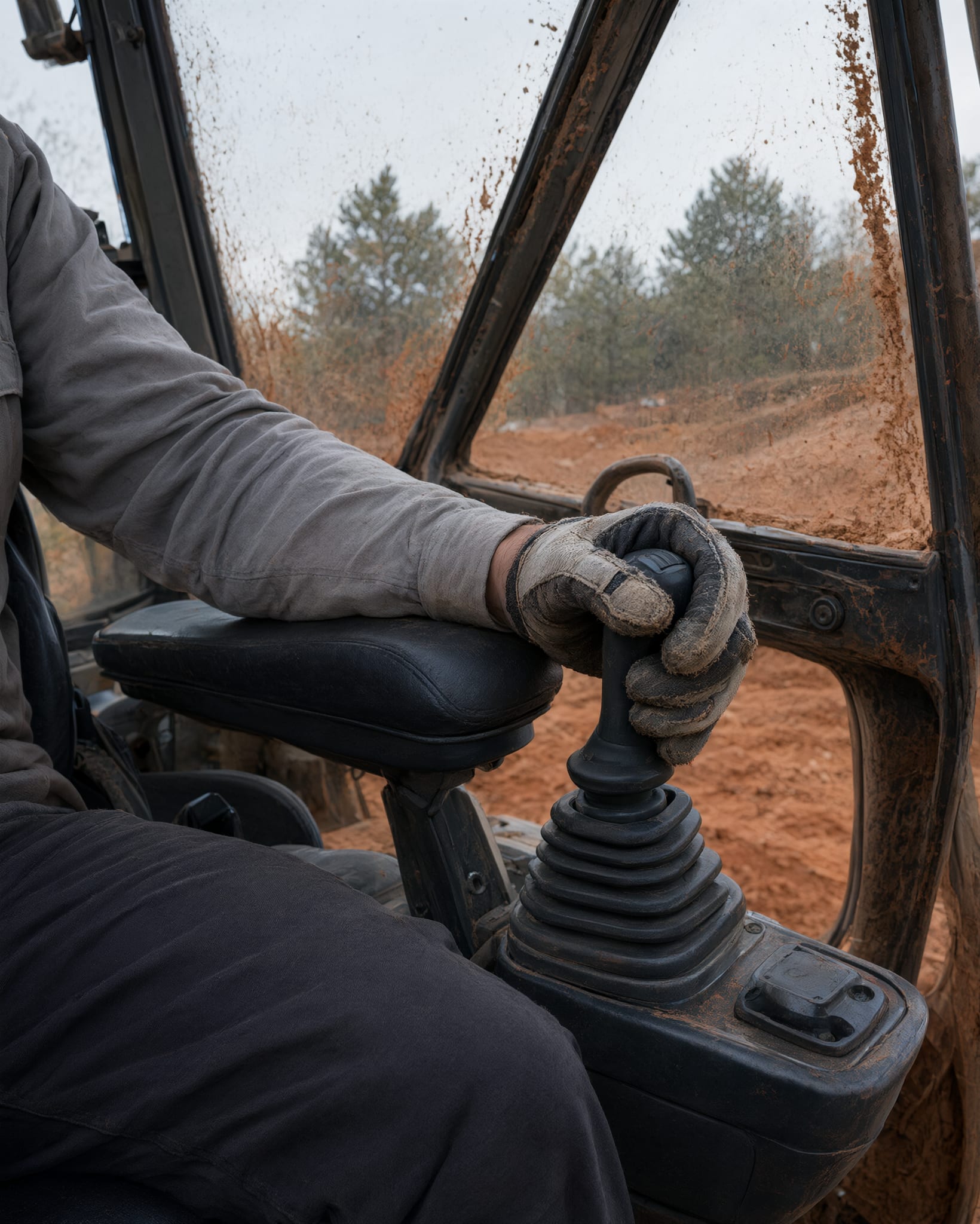 Operator's gloved hand on the right joystick of an excavator, forearm on the armrest, red clay splattered on the glass